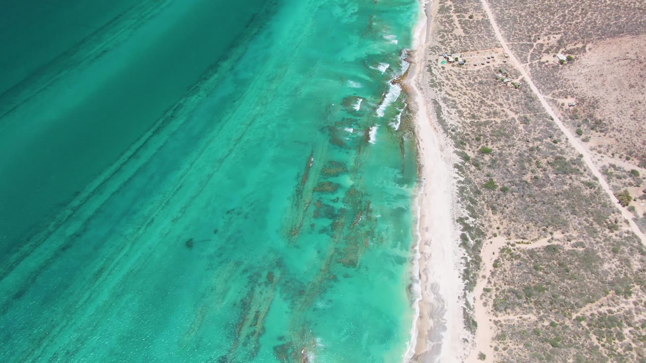 Cabo Pulmo aerial frame with shallow turquoise water, darker underwater bands, a thin sandy beach, dry vegetation, dirt tracks, and no visible people.