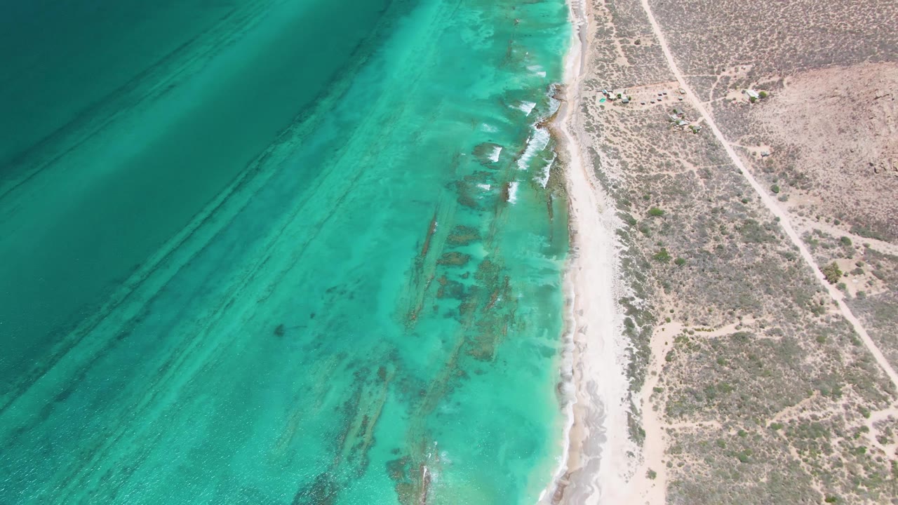 Mid-clip aerial view in Cabo Pulmo with turquoise ocean filling the left frame, small white wave lines along sand, arid scrubland to the right, and no visible people.