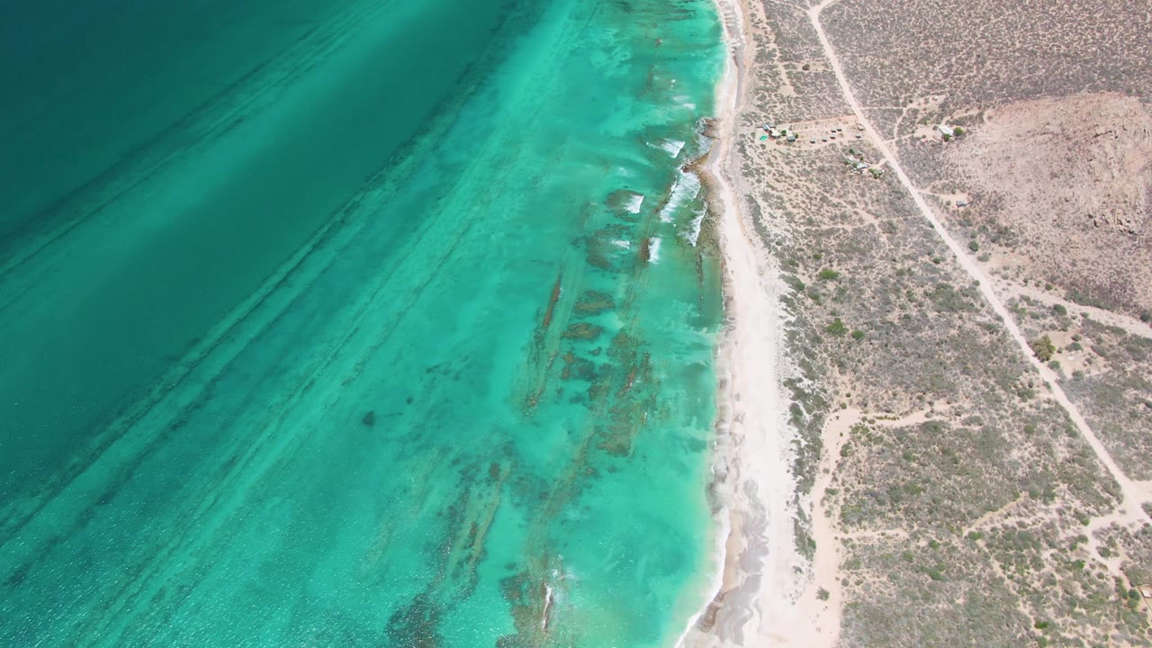 A later aerial frame over Cabo Pulmo where the turquoise water and darker underwater streaks lead into a bright sand strip, dry brush, dirt roads, and no visible people.