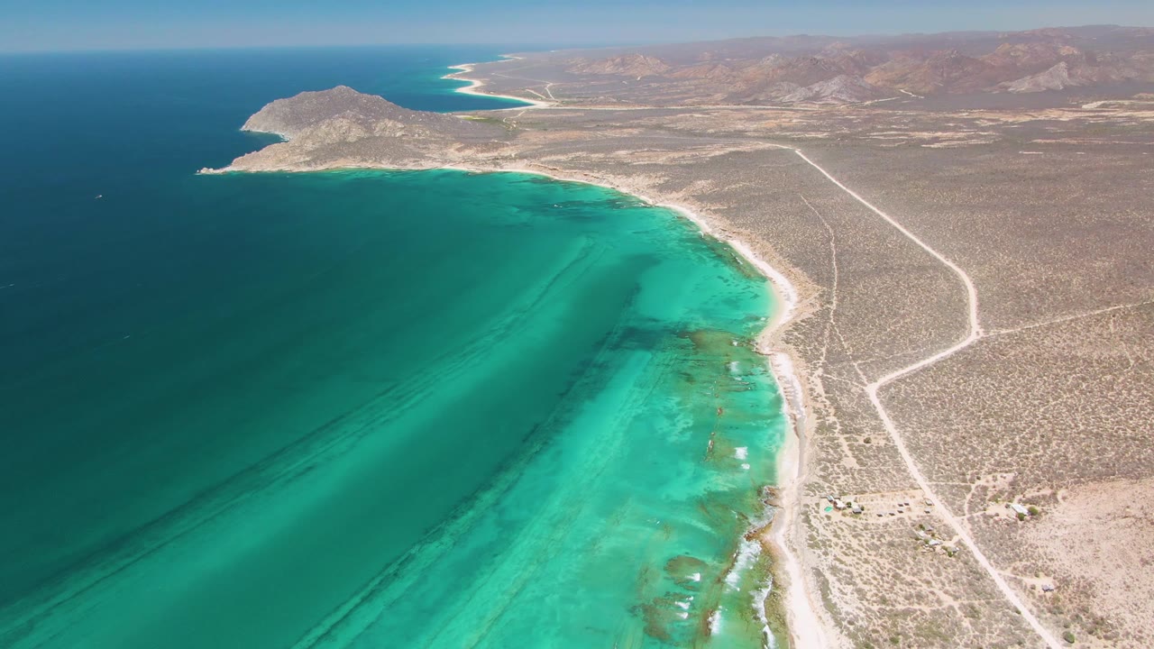 High aerial frame over Cabo Pulmo showing turquoise shallow water, pale beach, dry desert roads, distant hills, a small offshore boat, and no visible people.
