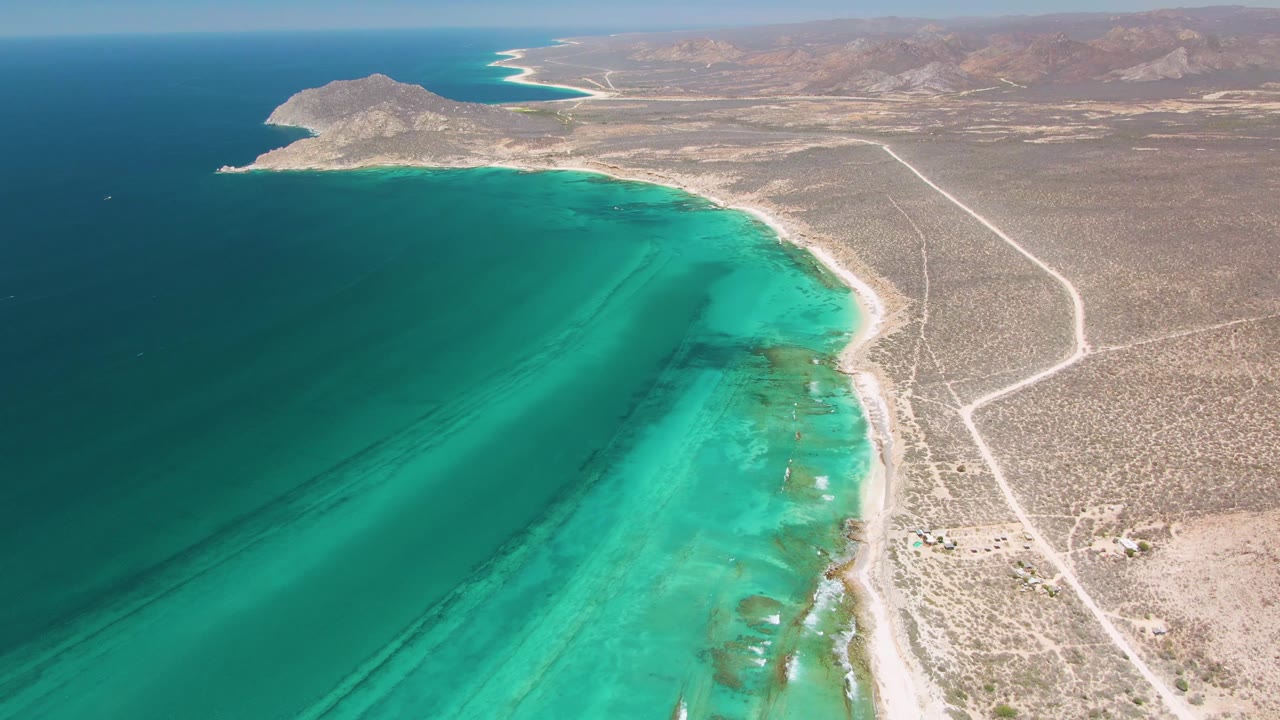 Aerial view in Cabo Pulmo with clear blue-green water filling the left side, a narrow sand shore, branching dirt roads inland, distant hills, and no visible people.