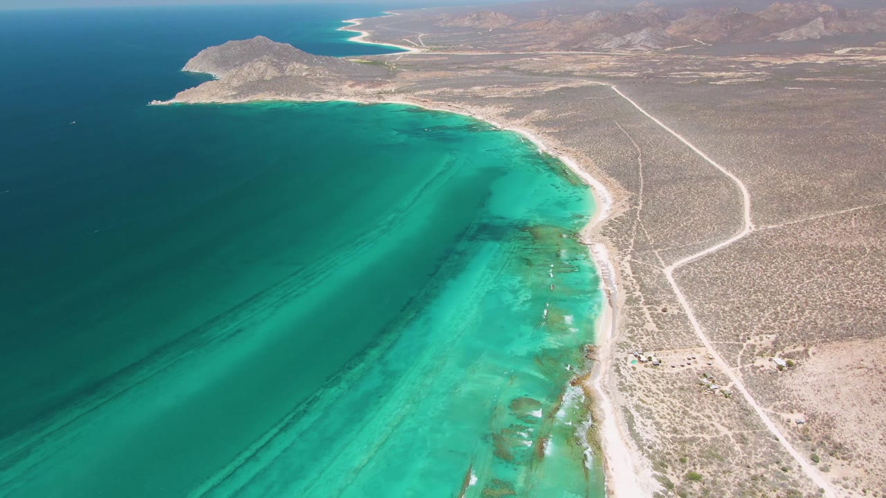 Forward aerial frame over Cabo Pulmo showing a curved turquoise bay, exposed light sand, scrubby arid terrain, visible road tracks, distant hills, and no visible people.