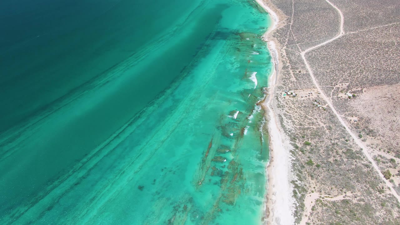 A second aerial frame of Cabo Pulmo shows the beach line shifted slightly through frame, with bright turquoise shallows, darker offshore water, scrub terrain, dirt tracks, and no visible people.