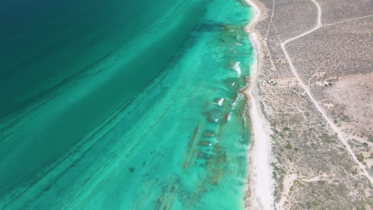 Aerial Cabo Pulmo coastline frame with more of the inland road network visible on the right, turquoise water on the left, a narrow beach between them, and no visible people.