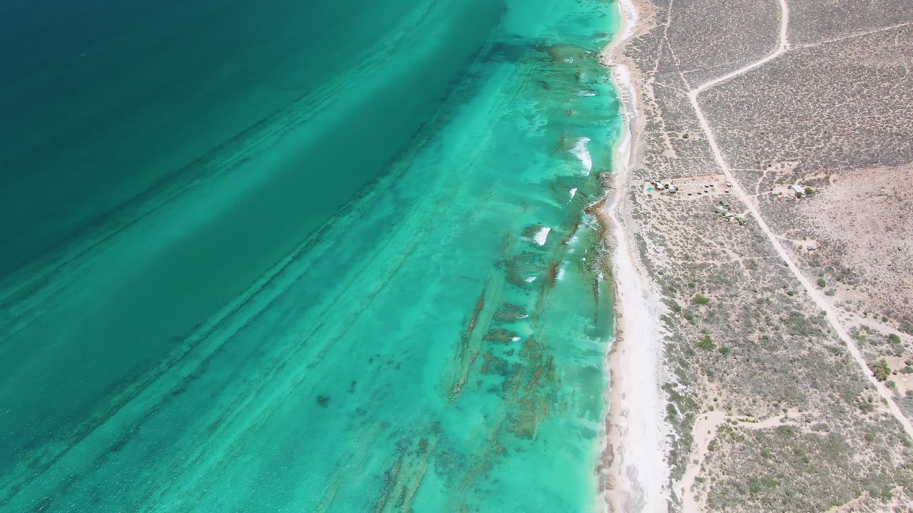 Mid-clip aerial frame over Cabo Pulmo showing calm turquoise water with darker underwater streaks, a pale beach edge, dry scrubland, dirt roads, and no visible people.