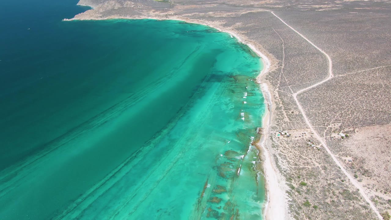 Aerial frame of Cabo Pulmo showing deep blue and turquoise coastal water beside a pale beach, dry scrubland, dirt roads, and no visible people.