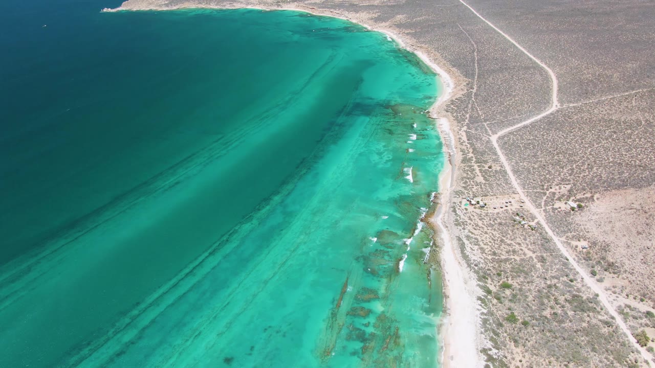 High drone view over Cabo Pulmo where small white surf breaks in turquoise shallows beside a narrow beach, with arid land and road tracks beyond and no visible people.