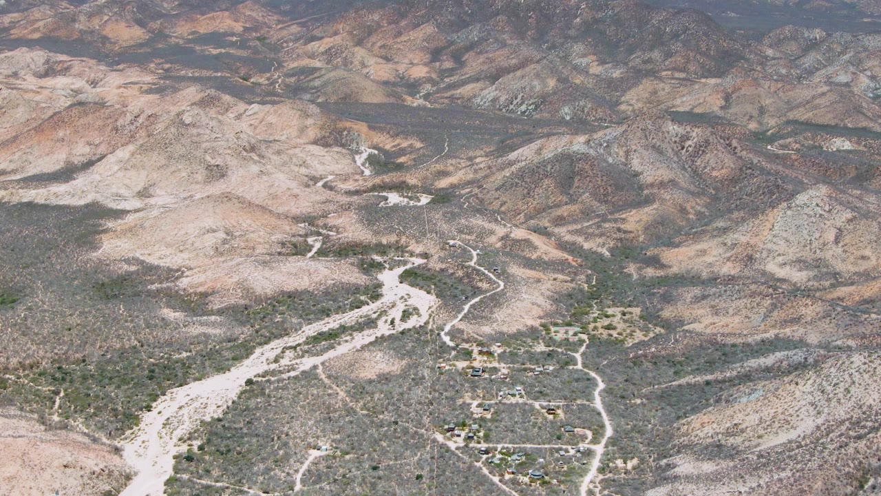 Aerial frame over Cabo Pulmo desert terrain, showing a pale dry wash, branching dirt roads, scrub-covered hills, distant mountains, and no visible people.