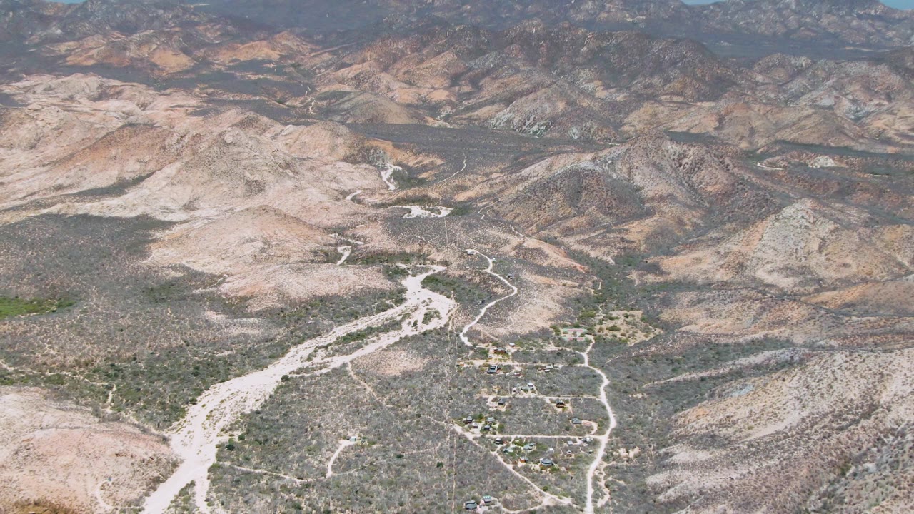 Cabo Pulmo aerial frame with a bright dry wash curving through rocky foothills, scattered vegetation, small buildings below, distant mountains, and no visible people.