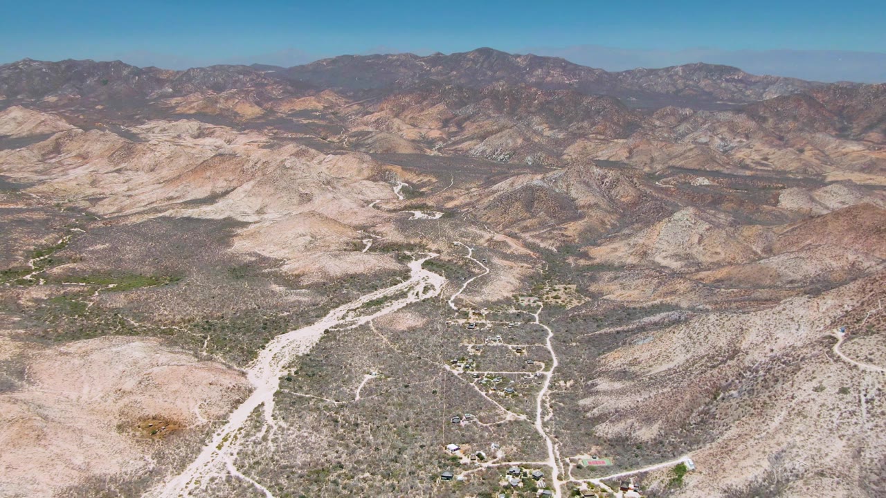 Wide aerial frame over Cabo Pulmo desert terrain, with pale dirt roads crossing scrub-covered hills toward layered mountain ranges under a clear blue sky and no visible people.
