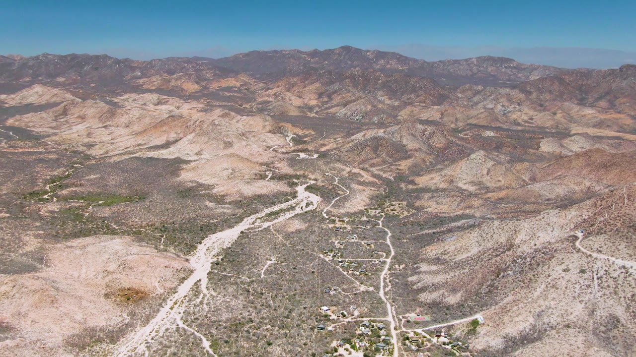High drone-style view near Cabo Pulmo showing a branching pale road in the desert foreground, low scrub across the valley floor, brown mountain ridges in the distance, and no visible people.