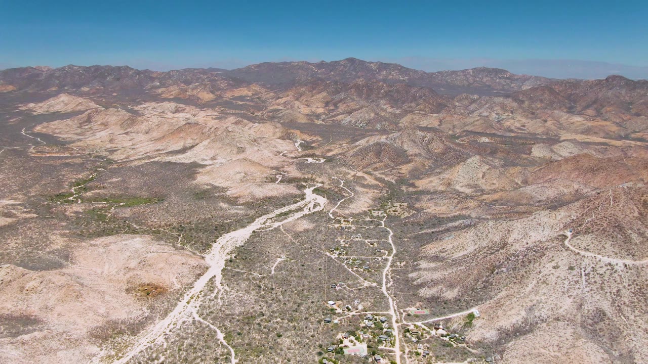 Aerial frame from Cabo Pulmo with a winding dirt road running through pale rocky terrain, sparse green shrubs, broad tan hills, distant mountains, and no visible people.
