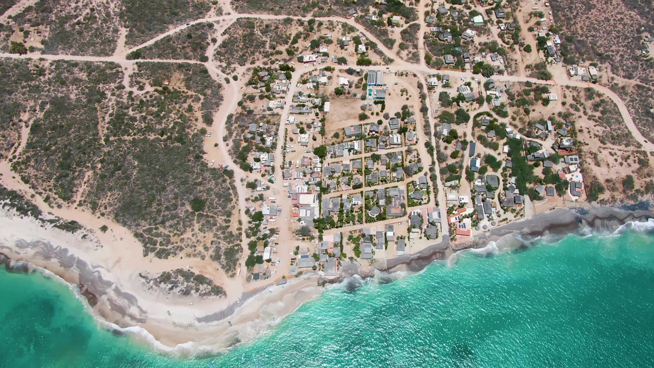 Aerial coastal frame with white waves breaking below a desert-edge neighborhood