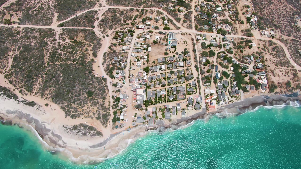 Overhead frame of Cabo Pulmo, Baja California Sur showing a small coastal neighborhood, pale dirt roads, sandy shore, turquoise water, and no visible people at the start of the clip.