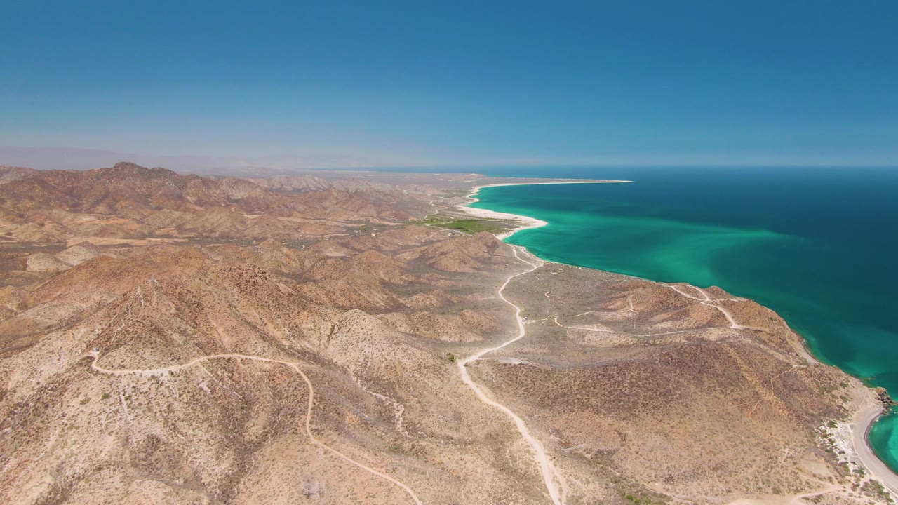 Opening aerial frame over Cabo Pulmo, Baja California Sur, with dry hills in the foreground, pale roads crossing toward a turquoise bay, and no visible people.