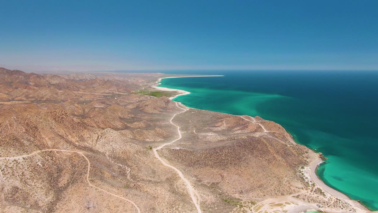 Second frame over Cabo Pulmo shows the desert foreground slightly shifted, with winding roads leading through rocky terrain toward the bright turquoise bay and no visible people.