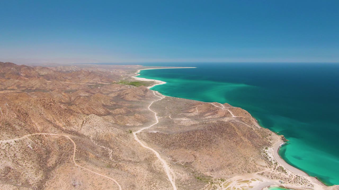 At 0:02, Cabo Pulmo appears from a broad drone angle, with ocher hills in the foreground, a branching dirt road, green-blue shallows near shore, deep blue water beyond, and no visible people.