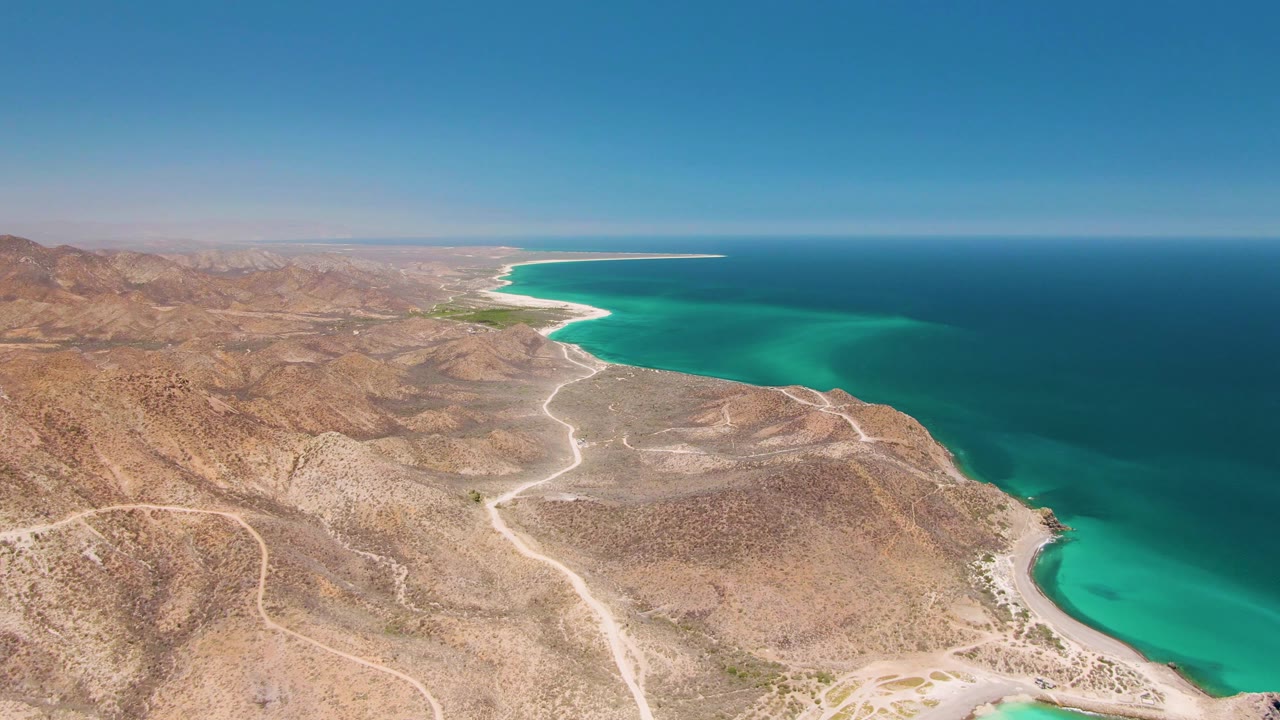 Mid-clip frame over Cabo Pulmo shows the shoreline lower in the composition, with dry ridges, pale tracks, turquoise water, a long ocean horizon, and no visible people.