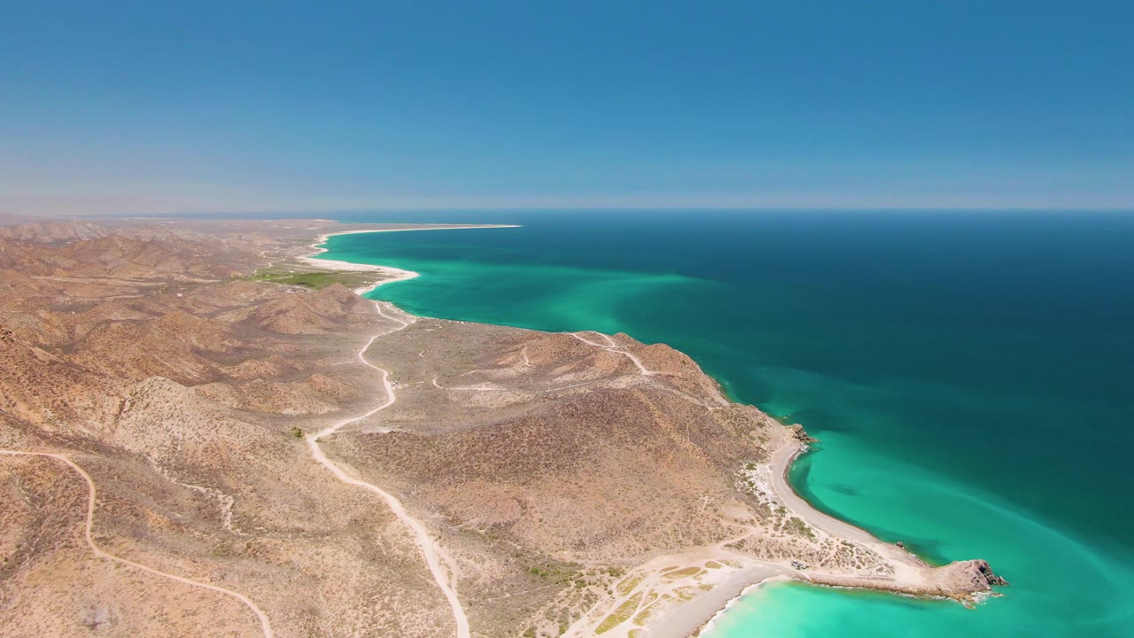 Aerial frame over Cabo Pulmo with tan desert hills, pale winding dirt roads, turquoise shallows, deep blue ocean, clear sky, and no visible people.