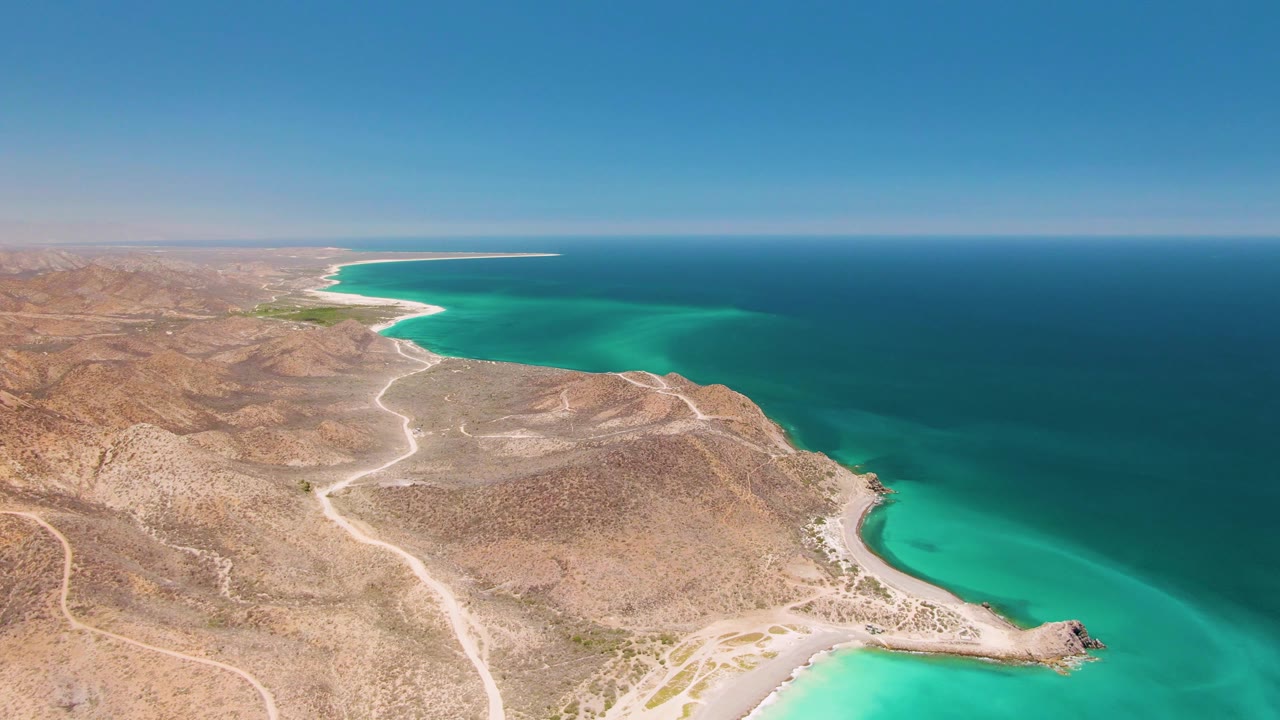 Aerial view of Cabo Pulmo with pale tracks crossing arid hills, a rocky point at the lower right, turquoise bay water, deep blue sea beyond, and no visible people.
