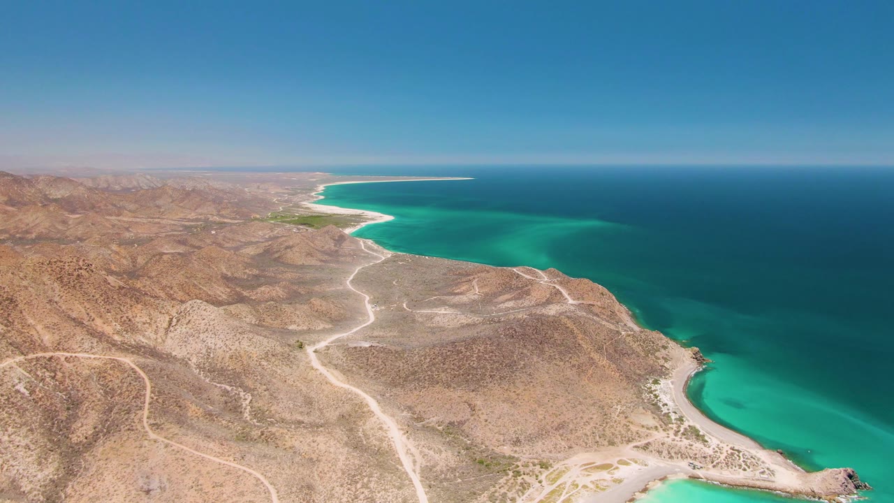 Opening aerial frame over Cabo Pulmo, Mexico, with tan desert hills, pale dirt tracks, turquoise cove water, deep blue ocean, clear sky, and no visible people.