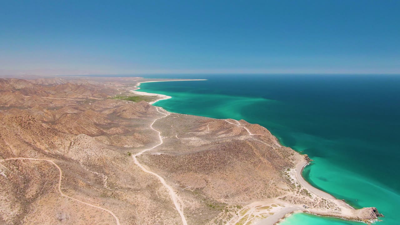 Aerial view one second into the Cabo Pulmo clip, showing the winding pale road across arid hills, a rocky point at lower right, turquoise shallows, deep ocean, and no visible people.
