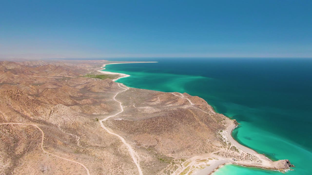 Aerial frame over Cabo Pulmo with the coastline stretching into the distance, the turquoise cove filling the right side, dry hills and dirt roads on the left, and no visible people.