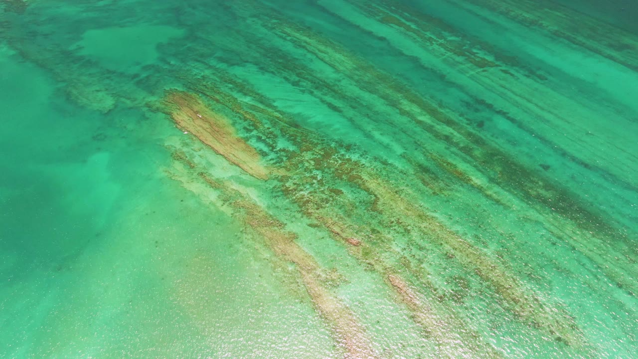 Top-down Cabo Pulmo ocean view with shallow turquoise water, elongated reef bands, and pale seabed texture visible through the surface, with no visible people.