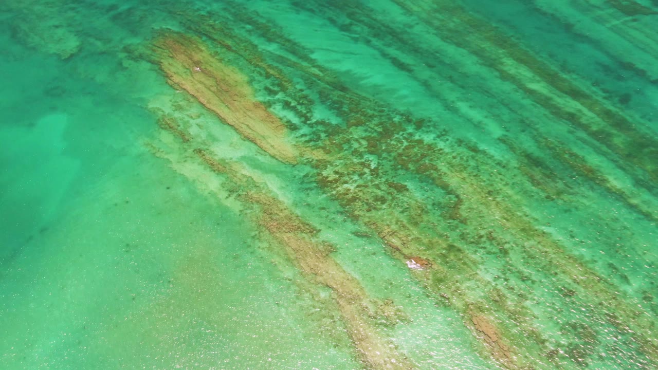 Overhead Cabo Pulmo frame where the submerged wooden structure angles through clear green-blue water beside darker ocean-floor bands, with no visible people.