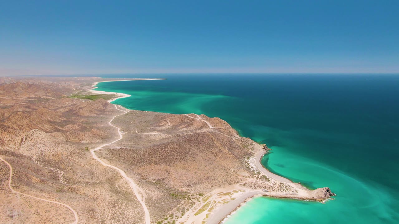 High aerial frame from Cabo Pulmo showing turquoise shallows along a rocky point, dry hills in the foreground, pale tracks across the terrain, and no visible people.