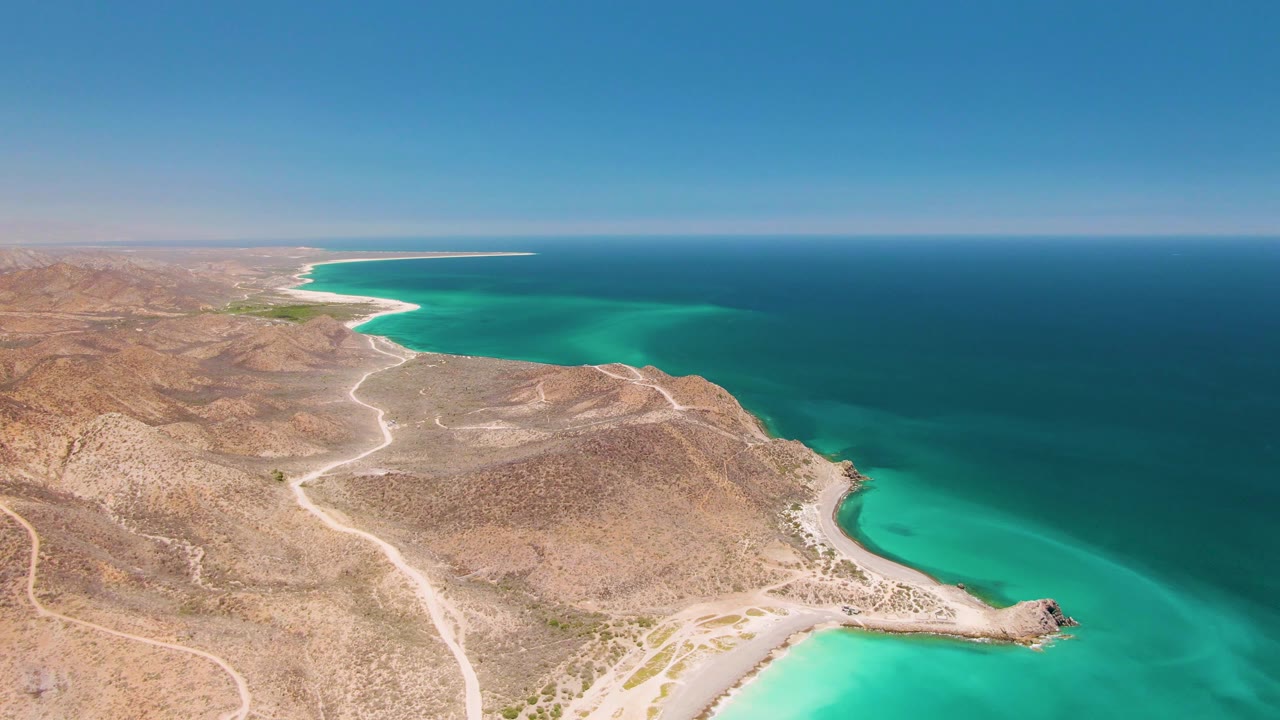 Drone-style aerial over Cabo Pulmo with arid tan ridges, branching dirt roads, bright green-blue nearshore water, deeper ocean beyond, and no visible people.