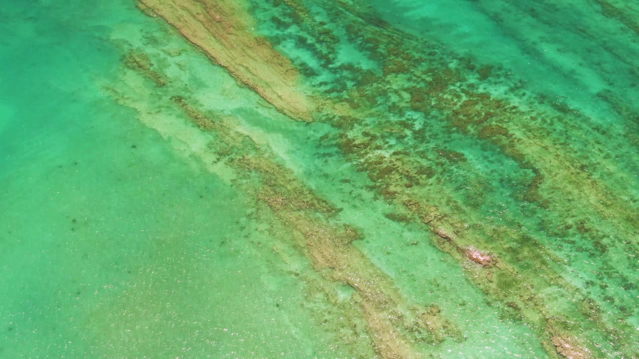 Overhead frame from Cabo Pulmo with broad turquoise water, diagonal tan underwater bands, green marine growth, faint surface ripples, and no visible people.