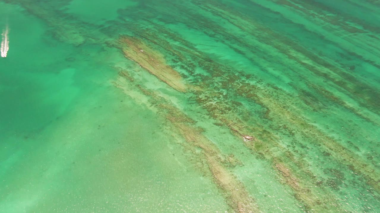 White sailboat crossing turquoise water above visible coral and sand patterns.