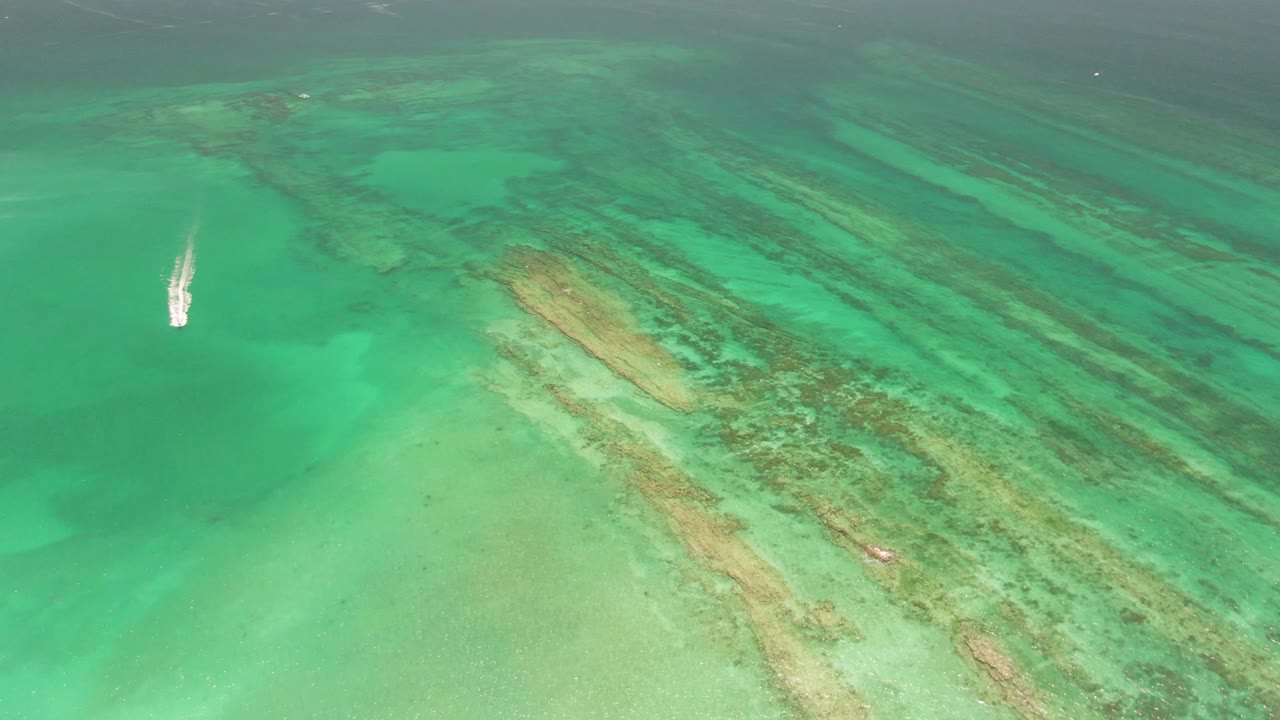 Aerial frame from Cabo Pulmo showing the small boat slightly farther across the turquoise water, with a white wake trailing beside darker reef streaks and no visible people.