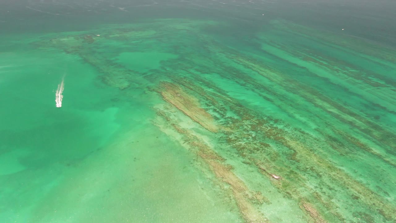 High-angle aerial view in Cabo Pulmo with the small boat near the upper-left water area, crossing clear green-blue sea above long diagonal coral and sand bands, with no visible people.