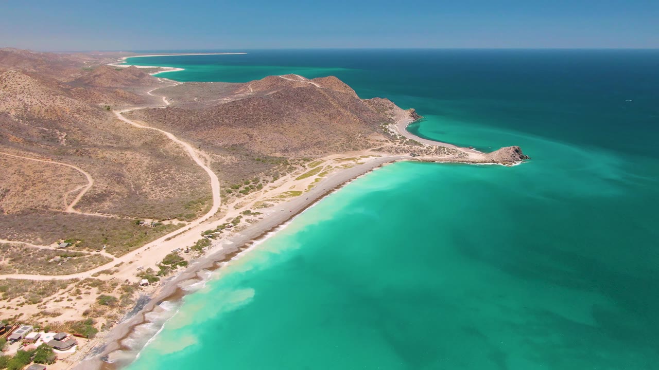Aerial view over Cabo Pulmo with turquoise water in the foreground, a pale sandy beach curving below rocky hills, dirt tracks inland, and no visible people.