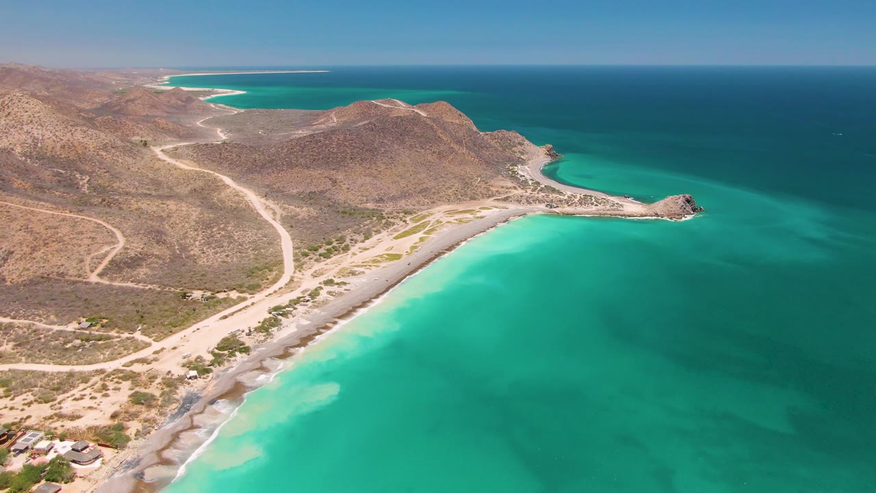 Turquoise bay water meets a pale beach below arid hills and dirt roads.