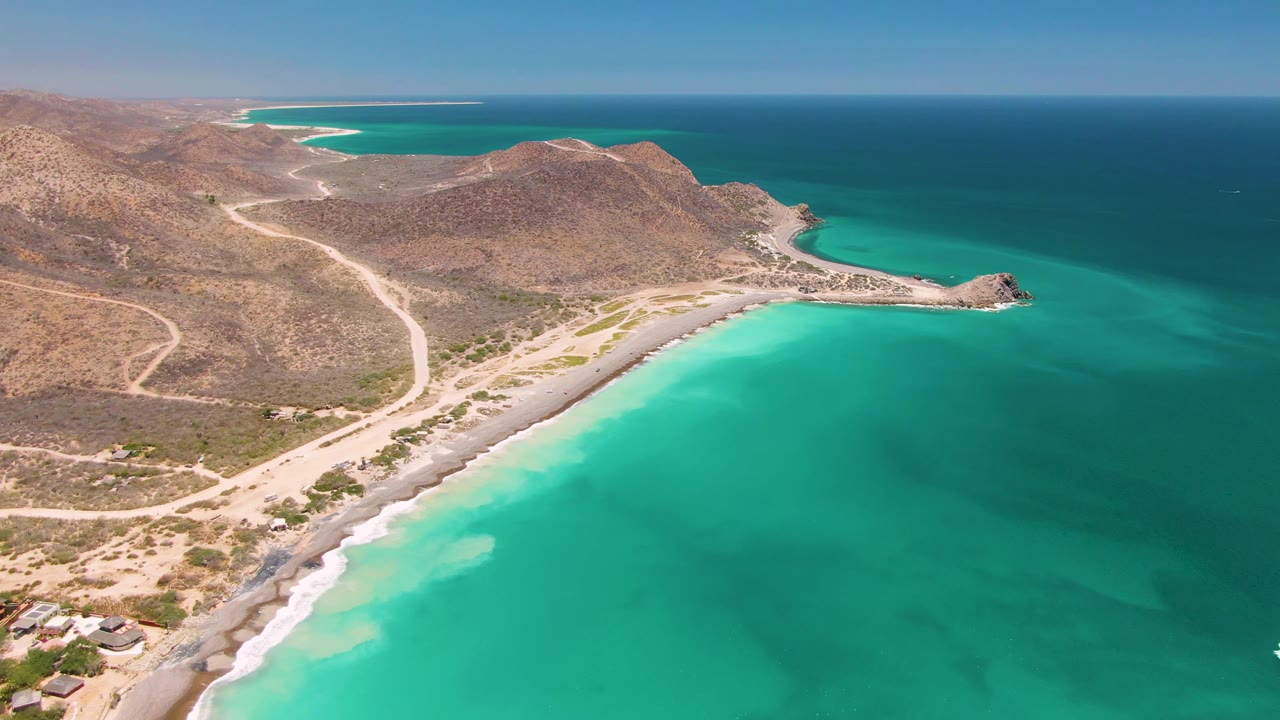 Aerial view over Cabo Pulmo showing clear turquoise water in the foreground, a sandy curved shore, arid hills, dirt tracks, and no visible people.