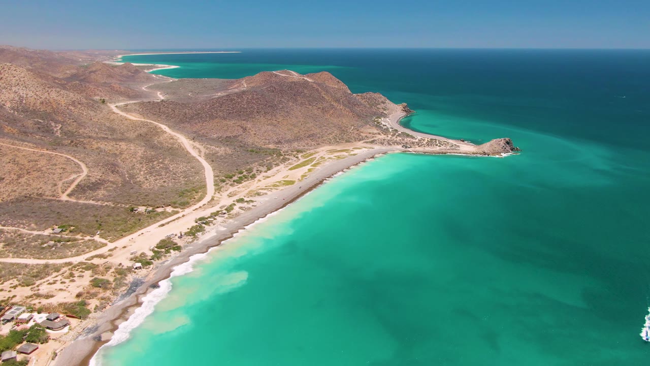 Wide aerial coastal view with turquoise water, dry hills, dirt roads, and sandy beach.