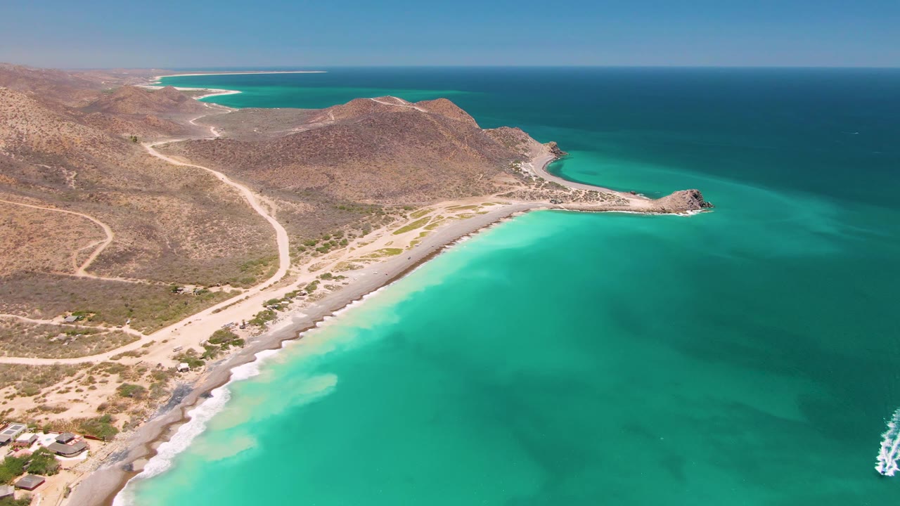 At two seconds, a white boat wake enters along the right edge of the Cabo Pulmo aerial view, beside clear turquoise water, sandy shore, arid hills, and no visible people.