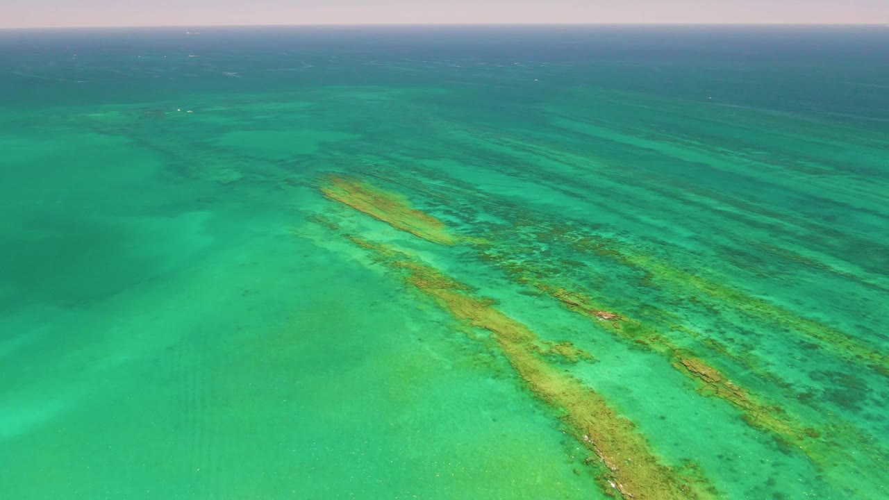 Aerial frame over Cabo Pulmo water with the reef line shifted slightly across the center, open sea at the horizon, and no visible people.