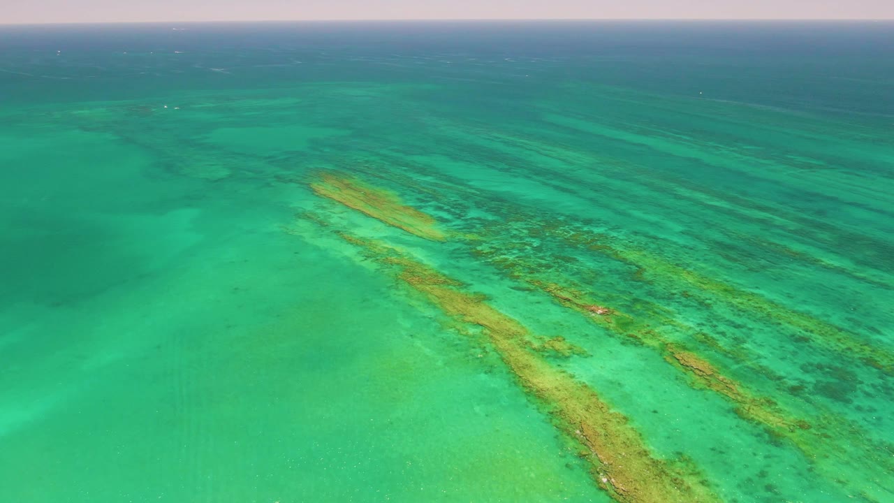 Mid-clip aerial view in Cabo Pulmo with diagonal reef patches clearer on the right side, pale shallow water in the foreground, and no visible people.