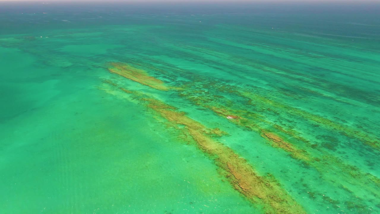 Aerial view of Cabo Pulmo clear shallow sea with elongated reef formations crossing the center of the frame and open water beyond, with no visible people.