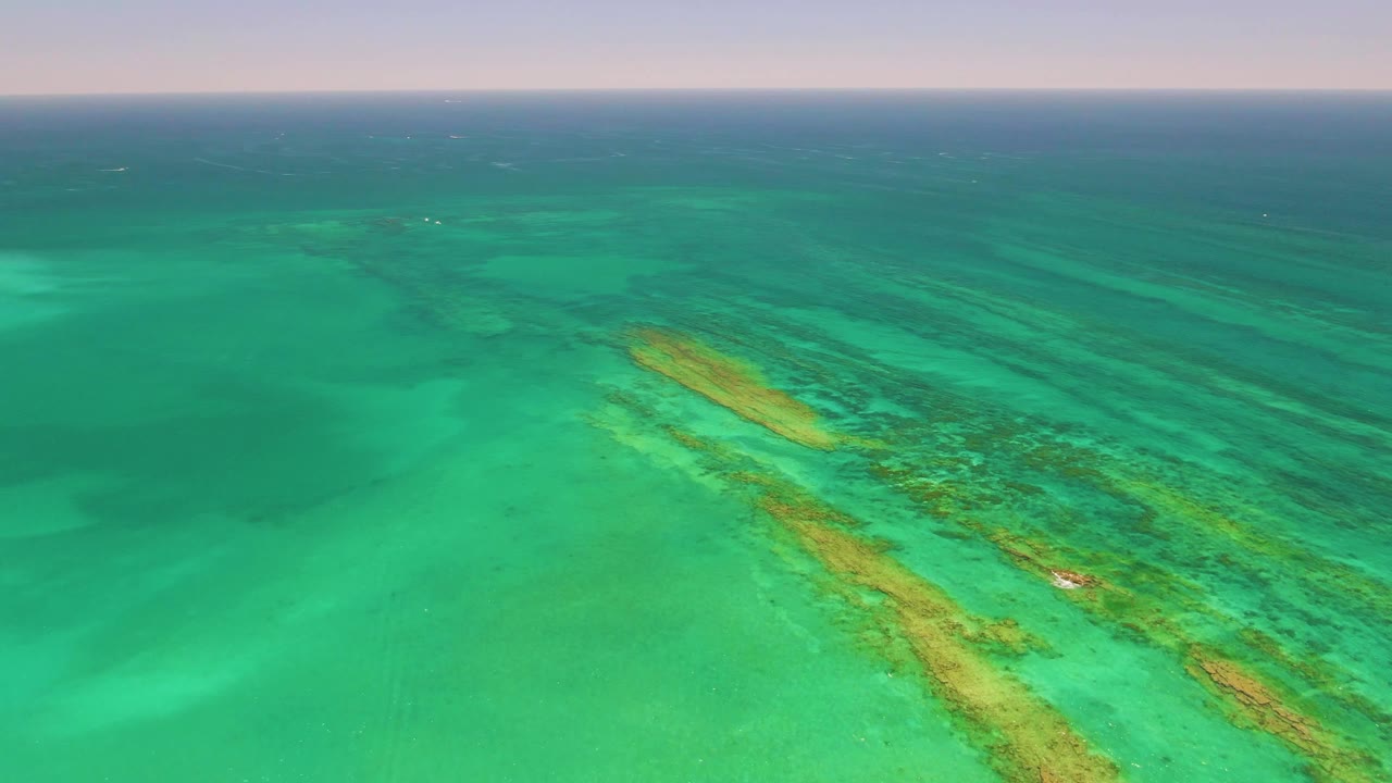 Aerial frame over Cabo Pulmo, Baja California Sur showing turquoise shallow water, submerged reef streaks, darker water near the horizon, and no visible people.