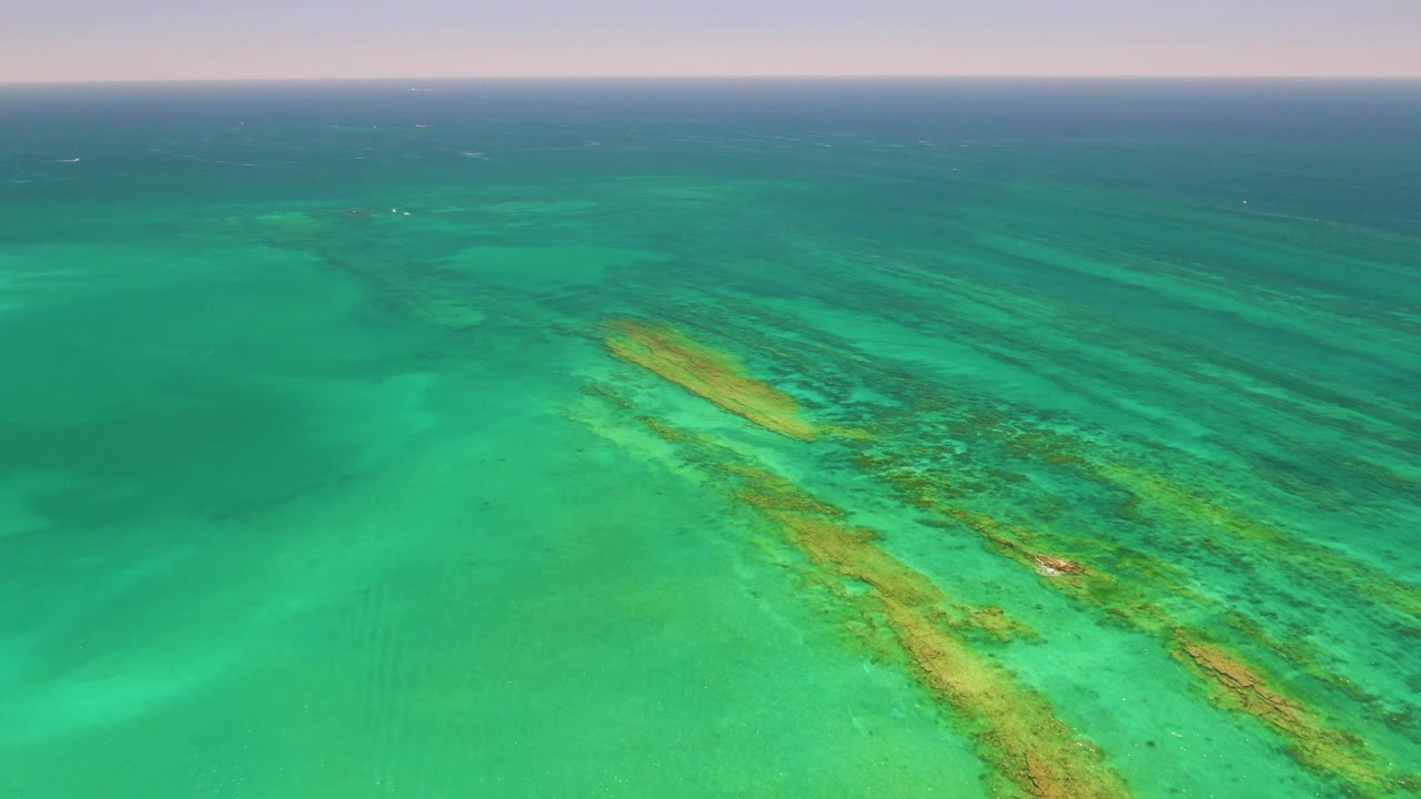Aerial frame over Cabo Pulmo, Baja California Sur showing diagonal reef formations under green-blue water, open sea in the background, and no visible people.