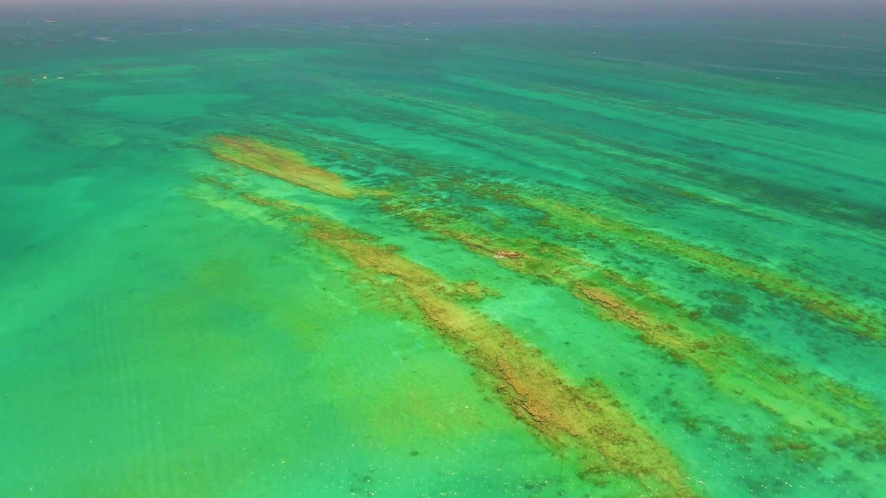 High aerial view over clear turquoise water in Cabo Pulmo, Mexico, showing tan underwater sandbars and darker coral patches with no visible people.
