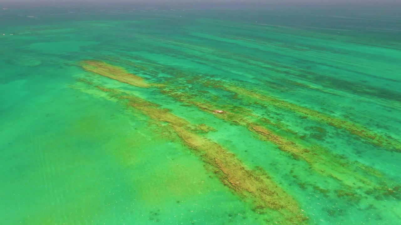 Oblique drone view in Cabo Pulmo, Mexico, with broad turquoise shallows, darker coral areas, and a small bright boat speck near the underwater formations, with no visible people.