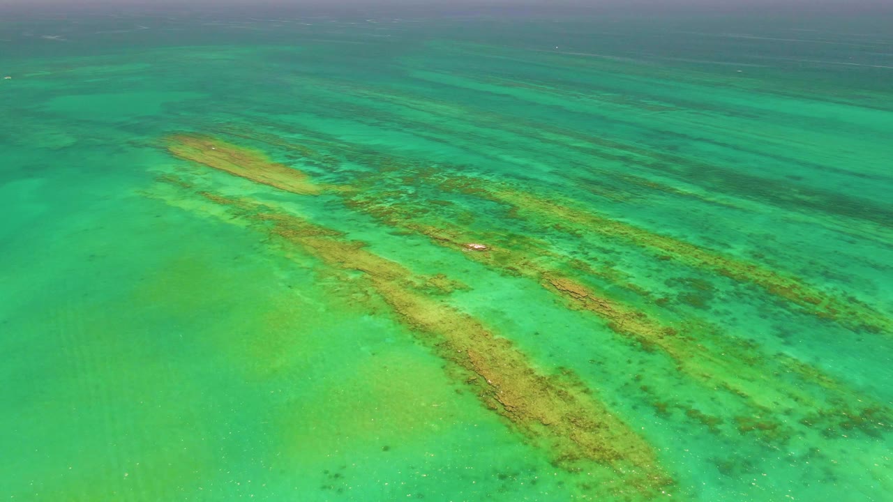 Aerial ocean view with sandbars, coral, and a tiny white boat.