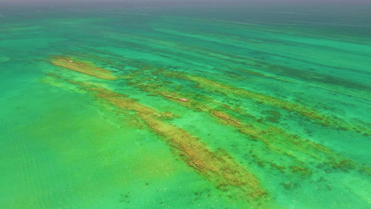 Overhead view of turquoise water in Cabo Pulmo with tan reef bands and darker underwater patches visible below ripples, no visible people.