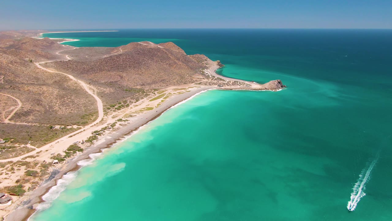 Aerial view of a boat wake in turquoise water beside a sandy coast and dry hills.
